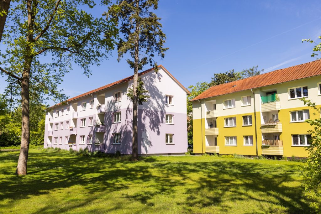 Mehrgeschossige Wohnhäuser mit gelber und heller Fassade, roten Ziegeldächern und Balkonen neben einer großen Rasenfläche mit Bäumen.